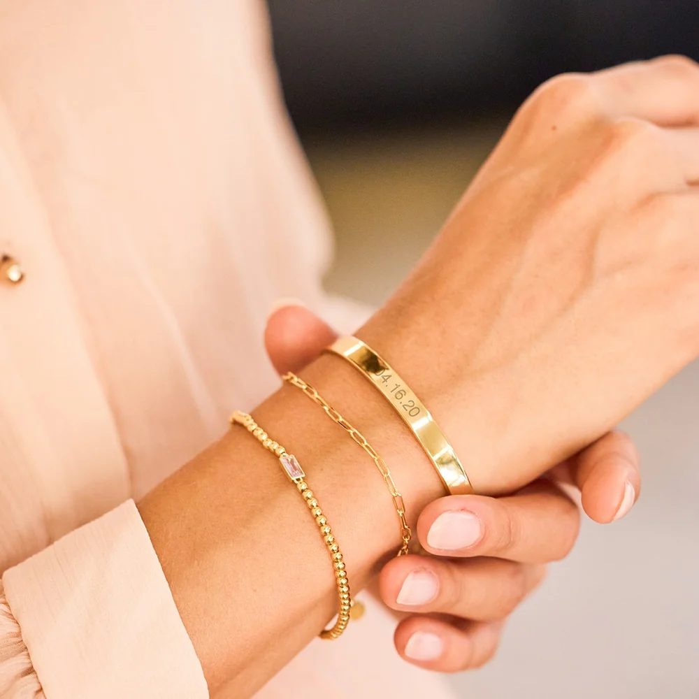 Brazalete rígido de oro con una fecha grabada, junto a dos pulseras finas